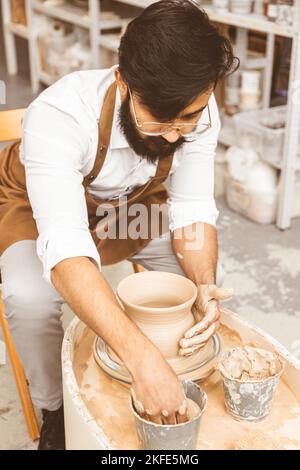 Ein junger männlicher Töpfer ist in seiner Werkstatt auf einem Töpferrad im Handwerk beschäftigt und stellt ein Tonprodukt her Stockfoto
