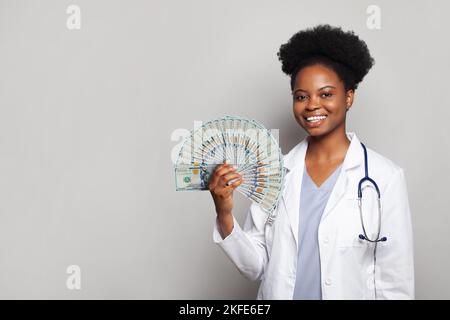 Erfolgreiche afroamerikanische Ärztin gp lächelt und hält vor grauem Hintergrund einen Geldfan von hundert Dollar fest Stockfoto