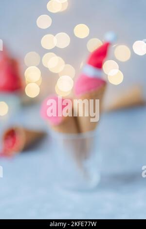 Defokussiert rosa Eis in Santa Hüte in einem Waffelkegel auf einem grauen Tisch, Hintergrund Girlanden Bokeh. Festliches gefrorenes Dessert Stockfoto