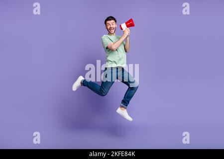Foto in voller Größe von schönen jungen Mann springen halten Megaphon aufgeregt gekleidet stilvolle graue Kleidungsstück auf lila Hintergrund isoliert Stockfoto