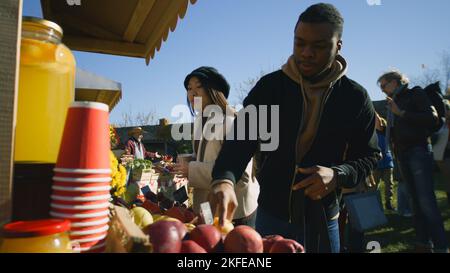 Vielfältiges Paar am Stand, das Obst und Gemüse auswählt. Menschen, die im Hintergrund spazieren und einkaufen. Lokaler Bauernmarkt oder Fair im Freien. Vegetarische und Bio-Lebensmittel. Landwirtschaft. Stockfoto