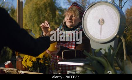 Ältere Verkäuferin steht am Stand und gibt Honig zum Geschmack. Die Menschen bezahlen für ein Glas Honig mit Bargeld. Lokaler Bauernmarkt oder Herbstmarkt im Freien. Vegetarische und Bio-Lebensmittel. Landwirtschaft. Stockfoto