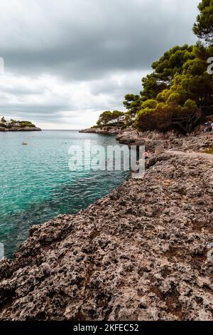 An einem bewölkten Tag steht ein vertikaler Felsstrand mit grünen Bäumen vor einem ruhigen Meer auf Mallorca Stockfoto