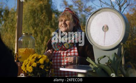 Ältere Verkäuferin steht am Stand und gibt Honig zum Geschmack. Die Menschen bezahlen für ein Glas Honig mit Bargeld. Lokaler Bauernmarkt oder Herbstmarkt im Freien. Vegetarische und Bio-Lebensmittel. Landwirtschaft. Stockfoto