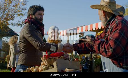 Ehepartner einkaufen auf dem lokalen Bauernmarkt. Frau steht mit Korb mit Obst oder Gemüse. Der Ehemann bezahlt den Kauf kontaktlos. Vegetarische und Bio-Lebensmittel. Landwirtschaft. Stockfoto