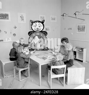 In der 1960s. Das örtliche Einkaufszentrum Farsta Centrum und die Gegend bieten Eltern-Käufern einen Service, bei dem sie beim Einkaufen nach ihren Kindern Ausschau halten. Im Bild vier Kinder mit einer Frau am Tisch beim Lesen und Spielen. Eine Lernuhr ist sichtbar, mit extra großen Zahlen für Stunden, mit denen die Kinder spielen und lernen können. Schweden 1967. Conard Ref. 5427 Stockfoto