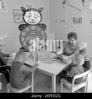 In der 1960s. Das örtliche Einkaufszentrum Farsta Centrum und die Gegend bieten Eltern-Käufern einen Service, bei dem sie beim Einkaufen nach ihren Kindern Ausschau halten. Im Bild vier Kinder mit einer Frau am Tisch beim Lesen und Spielen. Eine Lernuhr ist sichtbar, mit extra großen Zahlen für Stunden, mit denen die Kinder spielen und lernen können. Schweden 1967. Conard Ref. 5427 Stockfoto