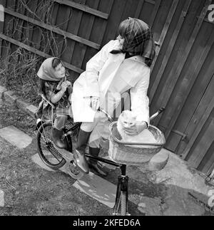 Eine Mutter im Alter von 1960s Jahren Eine junge lächelnde Frau ist in Regenkleidung gekleidet und fährt mit ihrer Tochter und einer Katze vor dem Fahrrad in einem Korb mit dem Fahrrad. Schweden 1967 Stockfoto