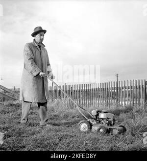 Gartengeräte der 1950s. Ein motorbetriebener Rasenmäher. Ein Mann wird gesehen, wie er den Rasen bewegt und das Gras schneidet. Er ist in der Freizeitkleidung des Jahrzehnts, in Mantel und Hut, sieht nicht zu glücklich aus. Schweden 1955 Stockfoto