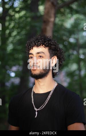 Ein männlicher Teenager mit lockigem Haar und einem Bart in einem schwarzen Hemd und ohne silbernen Hals im Wald Stockfoto