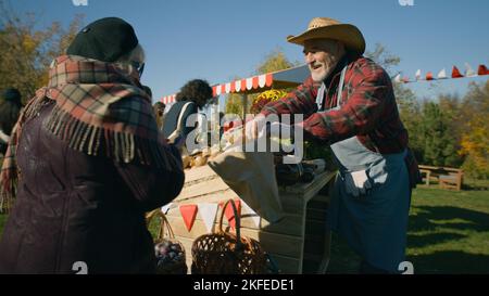 Ältere Frauen wählen am Stand Pflaumen mit Obst und Gemüse. Menschen, die im Hintergrund spazieren und einkaufen. Lokaler Bauernmarkt oder Fair im Freien. Vegetarische und Bio-Lebensmittel. Landwirtschaft. Stockfoto