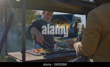 ZAPORIZHYA, UKRAINE - 01. NOVEMBER 2022: Menschen versuchen, Fleisch oder Gemüse auf dem Grill gekocht. Der Erwachsene Mann hebt bereit zum Essen ab und legt eine Portion auf den Teller. Wochenende auf dem lokalen Bauernmarkt. Stockfoto
