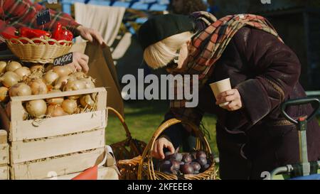 Ältere Frauen wählen am Stand Pflaumen mit Obst und Gemüse. Menschen, die im Hintergrund spazieren und einkaufen. Lokaler Bauernmarkt oder Herbstmarkt im Freien. Landwirtschaft. Point-of-Sale-System. Stockfoto