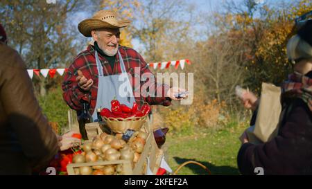 Seniorbauer gibt älteren Frauen Waren in Papiertüten. Frau zahlt für Einkäufe. Lokaler Bauernmarkt oder Herbstmarkt im Freien. Vegetarische und Bio-Lebensmittel. Landwirtschaft. Point-of-Sale-System. Stockfoto