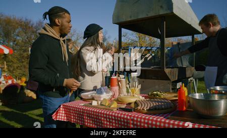 Multiethnisches Paar auf Herbstmesse oder Bauernmärkten. Asiatische Frau und afroamerikanischer Mann versuchen Käse. Mann kocht Fleisch auf dem Grill. Inhaber des POS-Systems. Picknick im Freien. Zeitlupe. Stockfoto