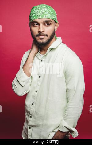Nachdenklicher junger indischer Mann in lässiger Kleidung mit Bandana auf dem Kopf und Blick vor rotem Hintergrund. Junge indische männliche Modell trägt Bandana. Stockfoto
