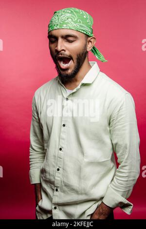 Gelangweilter junger indischer Mann, der eine Bandana auf dem Kopf trägt und mit einem geöffneten Mund auf rotem Hintergrund gähnt. Das Gähnen ist ansteckend, wenn man sich schlaf und schwach fühlt. Stockfoto