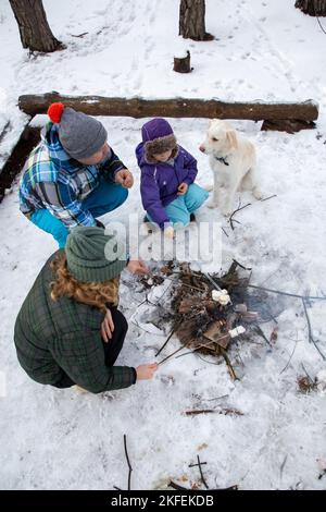 Familienpicknick im Winter. Glückliche Eltern, Kind, weißer Hund sitzen am Lagerfeuer im Wald und rösten während eines Winterwochenendes Marschlösser. Winter Stockfoto