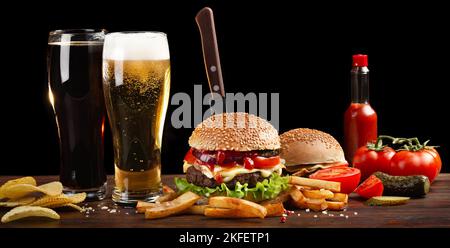 Hausgemachter Hamburger mit pommes Frites und zwei Gläsern Bier auf einem Holztisch. In den Burger steckte ein Messer. Fastfood auf dunklem Hintergrund. Stockfoto