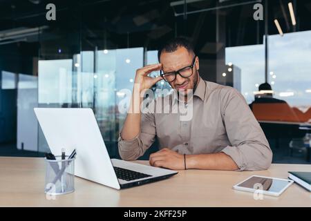 Kranker Mann bei der Arbeit mit einem Laptop, afroamerikanischer Mann mit starken Kopfschmerzen, Geschäftsmann, der im Büro arbeitet, trägt eine Brille. Stockfoto