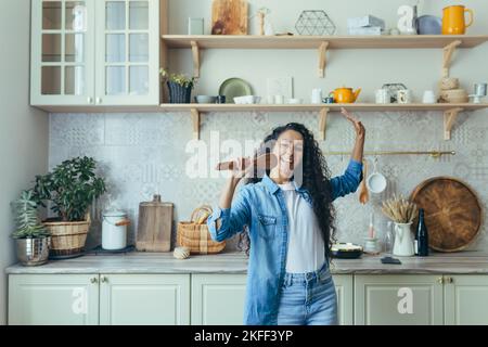 Lächelnde und glückliche junge schöne hispanische Frau in Denim-Kleidung, die in der Küche tanzt, einen Holzlöffel als Mikrofon hält und singt. Viel Spaß am Wochenende. Stockfoto