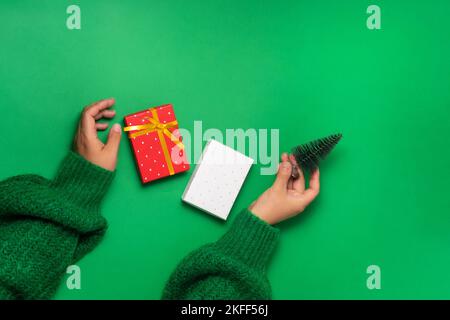 Frauenhände mit schwebender Geschenkschachtel und dekorativem weihnachtsbaum, Draufsicht auf grünem Hintergrund. Weihnachtsgeschenke Vorbereitungskonzept Stockfoto