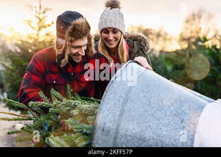 Glücklicher Mann und Frau verwenden Wickelvorrichtung mit Gitter, um einen Weihnachtsbaum zu verpacken, der auf dem weihnachtsmarkt gekauft wurde, um ihn nach Hause zu tragen Stockfoto