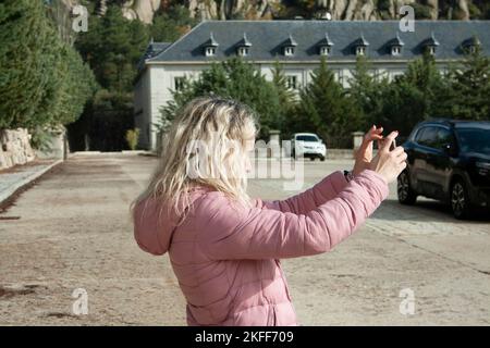 Blonde Frau, die mit dem Handy fotografiert, trägt an einem sonnigen Tag eine Winterjacke Stockfoto