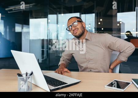 Überarbeiteter Geschäftsmann Rückenschmerzen, afroamerikanischer Mann massiert Rückenmann auf dem Stuhl sitzend mit Laptop bei der Arbeit, im modernen Bürogebäude. Stockfoto