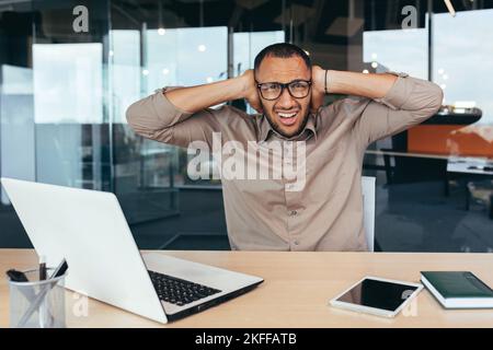 Besorgter Büroangestellter schaut auf die Kamera und bedeckt seine Ohren mit seinen Händen, der Lärm im Raum verhindert, dass der Arbeiter am Laptop im Gebäude arbeitet. Stockfoto