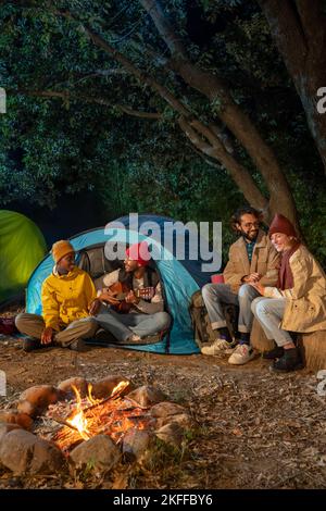 Glückliche Freunde, die Spaß zusammen am Lagerfeuer Camping spielen Gitarre in der Natur in der Nacht genießen. Stockfoto