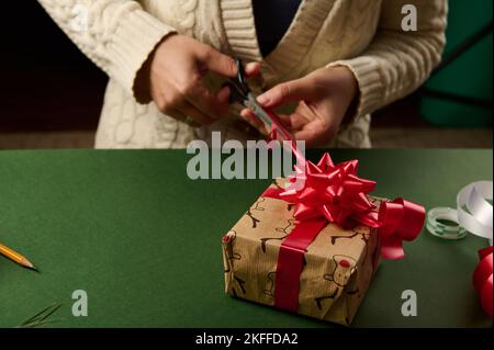 Nahaufnahme von Frauenhänden, die eine wunderschöne Schleife aus einem glänzenden roten Band binden, während sie ein Weihnachtsgeschenk dekorieren. Umhüllung. Stockfoto