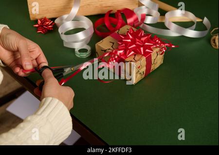 Nahaufnahme einer Frau, die ein glänzendes Band in Streifen schneidet, um wunderschöne Ornamente zu machen, während sie Geschenke zu Weihnachten einwickelt Stockfoto