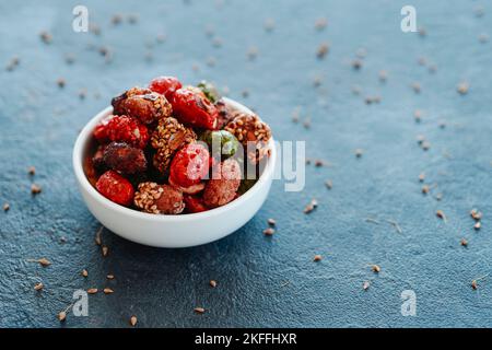 Eine Auswahl kandierter Erdnüsse, mit verschiedenfarbigen Zucker- und Samenbezügen, serviert in einer weißen Keramikschale mit, auf einer strukturierten, dunkelgrauen Steinbrandung Stockfoto