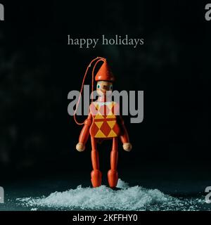 Der Text Happy Holidays und ein weihnachtsbaum-Ornament in Form einer Harlekin-Marionette auf einem Schneehaufen vor schwarzem Hintergrund Stockfoto