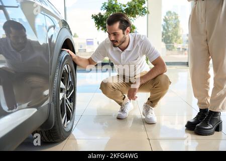 Lächelnder Autokäufer, der den Zustand der Autoreifen im Ausstellungsraum überprüft Stockfoto