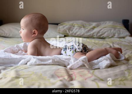 Ein glückliches, zufriedenes Baby, das auf dem Bauch liegt und in der Bauchlage sitzt, um ihren Rücken zu stärken. Sie trägt eine moderne, wiederverwendbare Stoffwindel Stockfoto