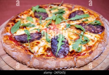 Frische Pizza aus dem Ofen mit Salami und Rucola auf einem Holztablett Stockfoto