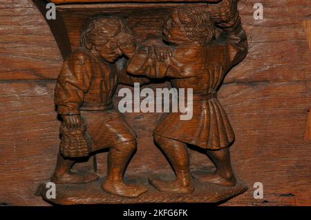 Zwei Männer kämpfen, aber während ein Mann bereit scheint, mit der rechten Hand zu schlagen, beruhigt oder beruhigt er die Stirn oder Braue seines Gegners mit der linken. In Holz geschnitzt um 1480 auf einem Misericord (Unterseite des Kippsitzes) in einem der Chorstände in der Oude Kerk (Alte Kirche) in Oudekerksplein, Amsterdam, Niederlande. Die Schnitzerei veranschaulicht die tödliche Sünde des Zorns oder Zorns im Gegensatz zur himmlischen Tugend der Güte oder Liebe und wurde möglicherweise von einem alten niederländischen Sprichwort inspiriert: „Einer greift an, während der andere beruhigt“. Stockfoto