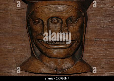 Kapuzengesicht mit zerklüfteten Zähnen, drei Augen und zwei Nasen, aus Holz geschnitzt, Ende 1400 n. Chr.. Misericord auf der Unterseite des Chorstuhlsitzes in der Oude Kerk (Alte Kirche), Amsterdam, Niederlande. Es wird vermutet, dass es sich um einen Kommentar handelt, wie betrunkene Menschen dazu neigen, sich über alles einig zu sein. Stockfoto