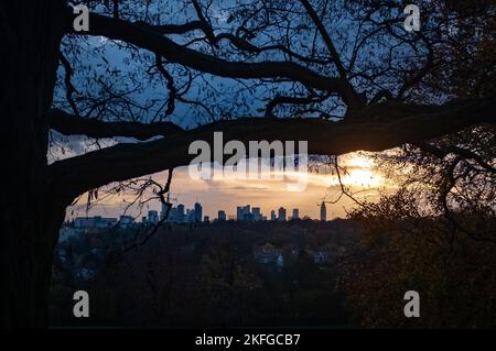 18. November 2022, Hessen, Frankfurt/Main: Die Sonne geht hinter der Frankfurter Wolkenkratzer-Kulisse unter, während im Vordergrund Bäume und Äste auf dem Lohrberg im Nordosten der Stadt zu sehen sind. Foto: Frank Rumpenhorst/dpa Stockfoto