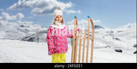 Kind in Winterkleidung, das mit einem Holzschlitten auf einem verschneiten Berg steht Stockfoto