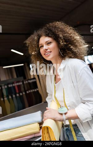 Erfreuliche Verkäuferin mit Nadelkissen auf der Hand, die gelbe Stoffrolle im Textilgeschäft hält Stockfoto