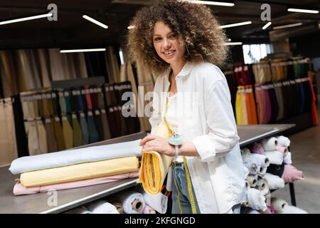 Fröhliche Verkäuferin mit Nadelkissen auf der Hand, die gelbe Stoffrolle im Textilgeschäft hält Stockfoto