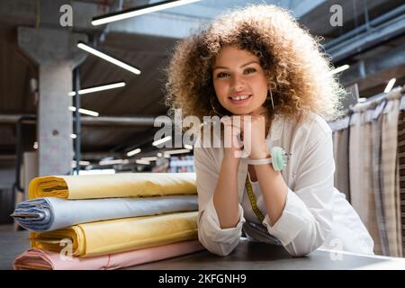 Eine erfreuende Verkäuferin mit Nadelkissen auf der Hand, die die Kamera in der Nähe von pastellfarbenen Stoffrollen ansah Stockfoto