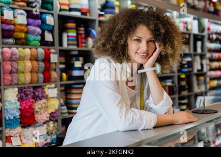 Junge Verkäuferin mit Nadelkissen auf der Hand, die in der Nähe der Theke und der Handarbeitszubehör die Kamera anlächelt Stockfoto