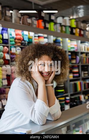 Glückliche und lockige Verkäuferin, die die Hände in der Nähe des Gesichts hält, während sie im Textilgeschäft die Kamera anschaut Stockfoto