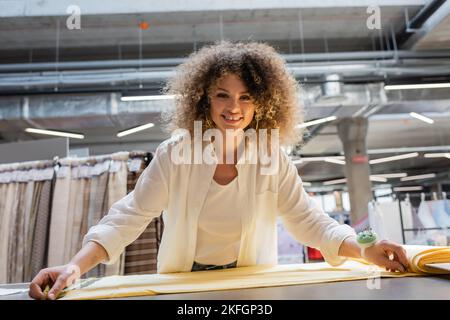 Fröhliche Verkäuferin mit Nadelkissen an der Hand, die gelben Stoff misst, im Textilgeschäft Stockfoto
