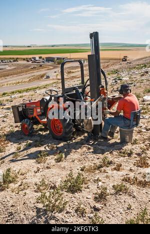 Ein männlicher Arbeiter, der Handschuhe und Gehörschutz trägt, arbeitet mit Gewinderohren, während er auf einem Traktor auf einer aktiven Deponie unterwegs ist. Stockfoto