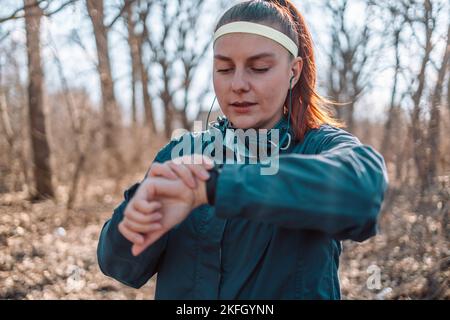 Sportuhr laufen Frau Überprüfung Smartwatch Tracker. Trail Running Läufer Mädchen Blick auf Herzfrequenzmesser Smart Uhr in Wald tragen Jacke Stockfoto